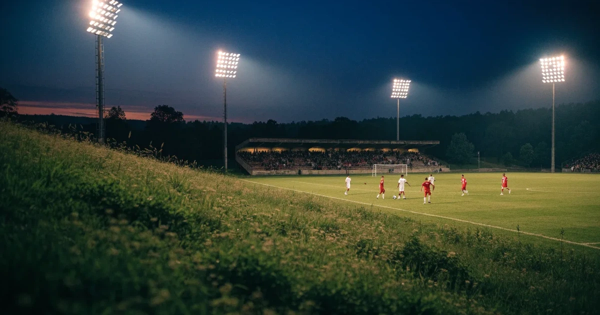 Ein Fußballstadion unter Flutlicht mit hellen Rängen, von einem Hügel aus gesehen, darüber ein dunkelblauer Abendhimmel.