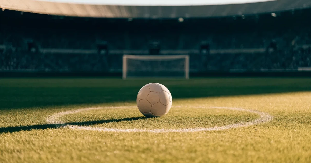 Ein Fußball liegt auf dem Anstoßpunkt eines leeren Rasenplatzes, im Hintergrund eine große Stadiontribüne im Abendlicht.
