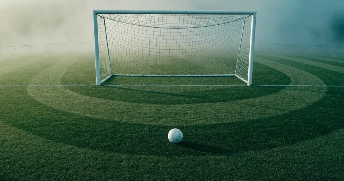 Ein Fußballtor mit gespanntem Netz steht auf einem leeren Rasenplatz, davor ein einzelner Ball, Morgendunst liegt über dem Feld.