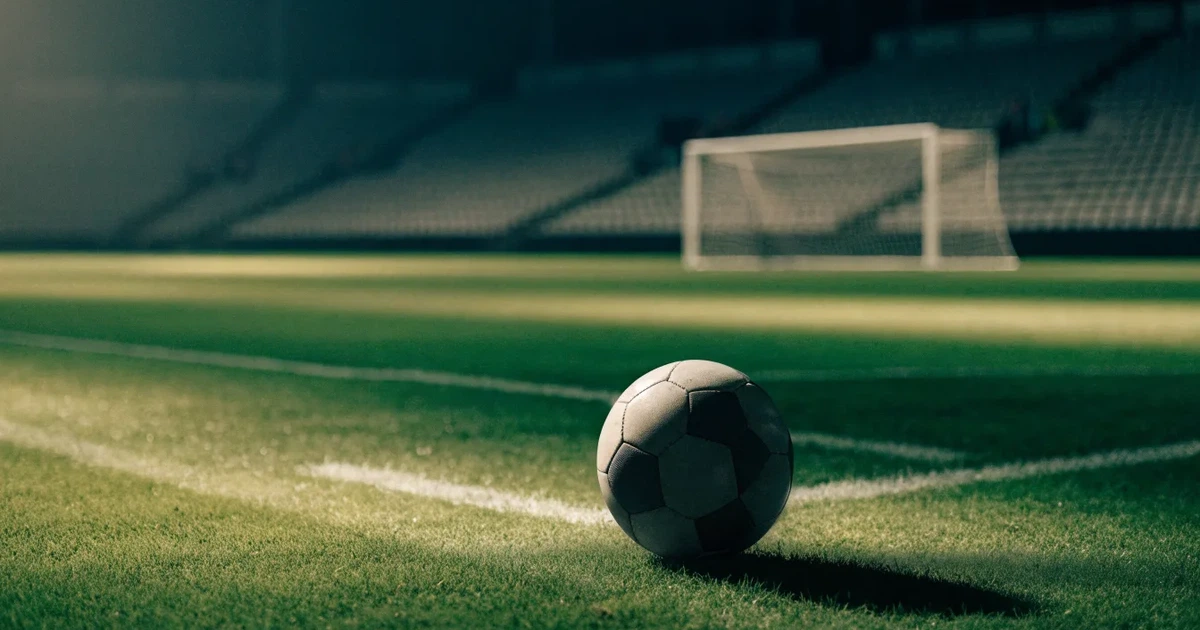Ein Fußball liegt auf dem Rasen eines leeren Stadions bei Flutlicht, im Hintergrund unscharfe Tribünen ohne erkennbare Beschriftung.