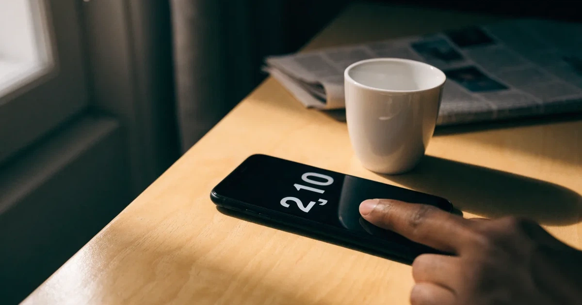 Ein Daumen schwebt über einem Smartphone mit dunklem Display, das auf einem Küchentisch neben einer Tasse liegt.