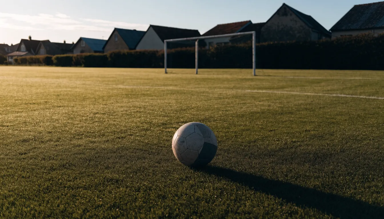 Ein Fußball auf dem Rasen eines leeren Sportplatzes am Rand einer Kleinstadt im Morgenlicht.
