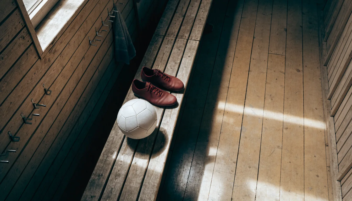 Ein Fußball liegt auf einer Holzbank in einer leeren Umkleide neben einem Paar Fußballschuhen ohne Markenzeichen.
