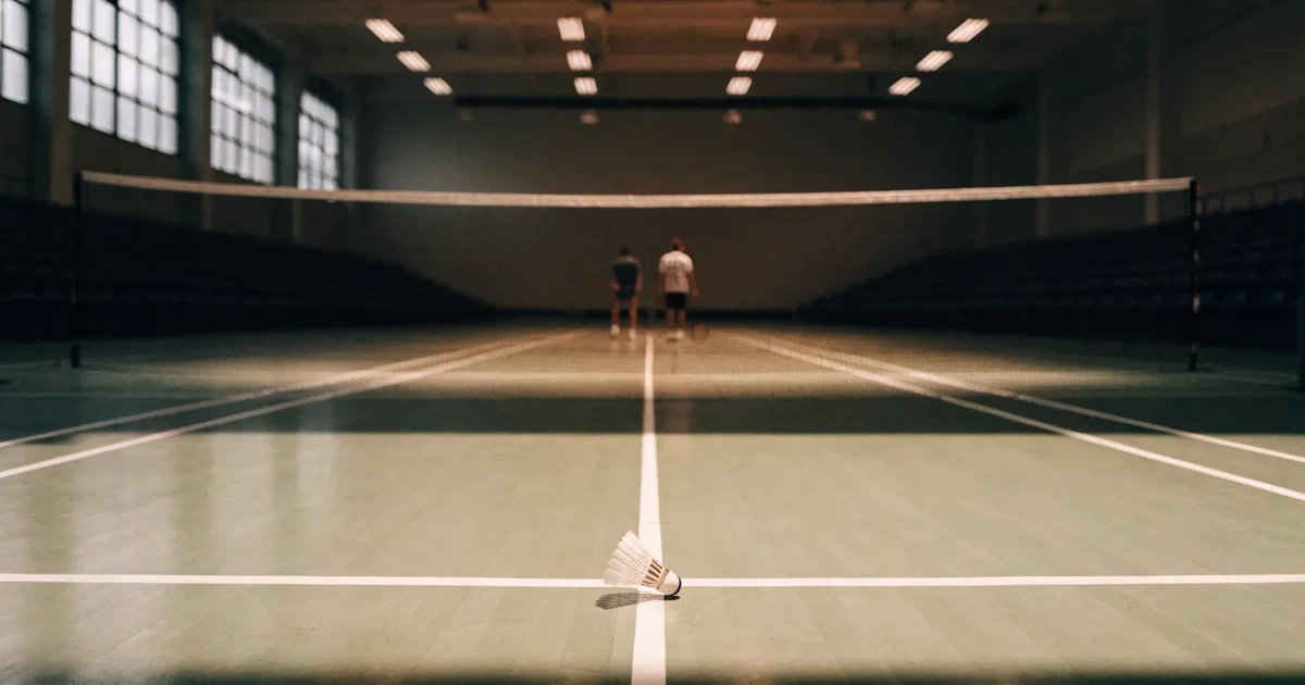 Eine leere Badmintonhalle mit gespanntem Netz und hellen Hallenlichtern, ein Federball liegt auf dem Hallenboden.