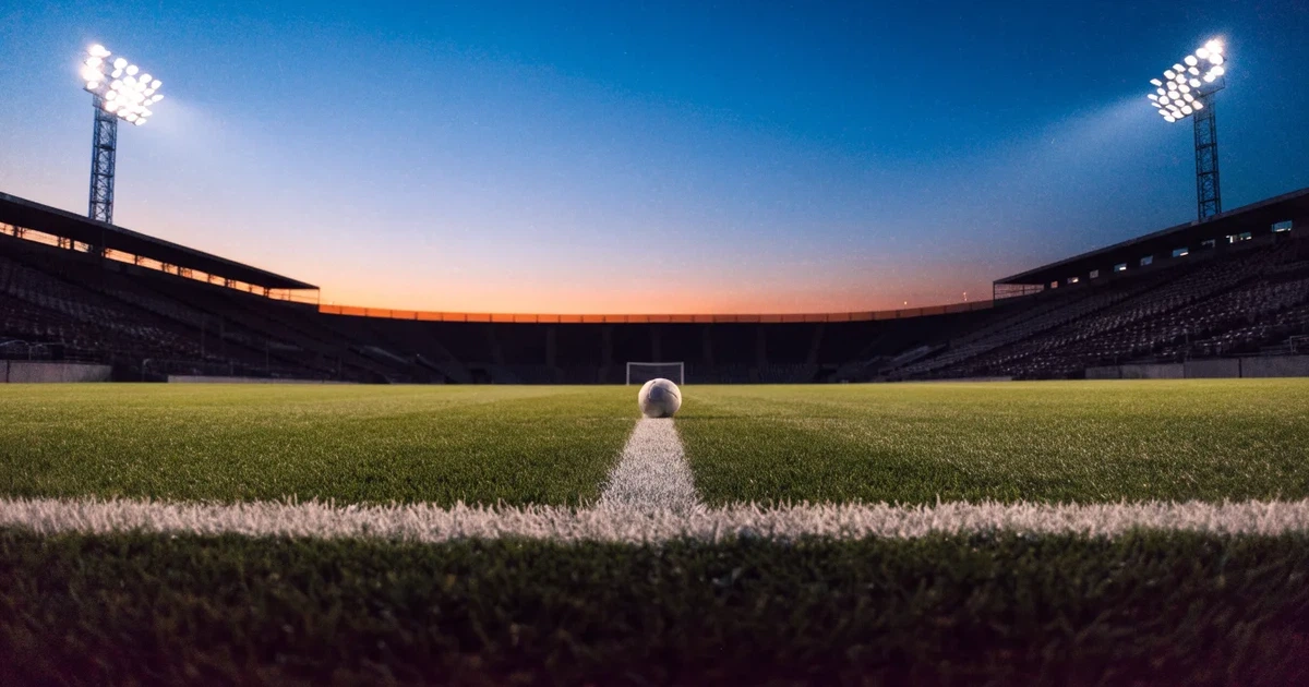 Ein Rasenplatz in der Dämmerung mit eingeschalteten Flutlichtmasten, ein Ball liegt am Anstoßpunkt, die Tribünen im Hintergrund sind unscharf.
