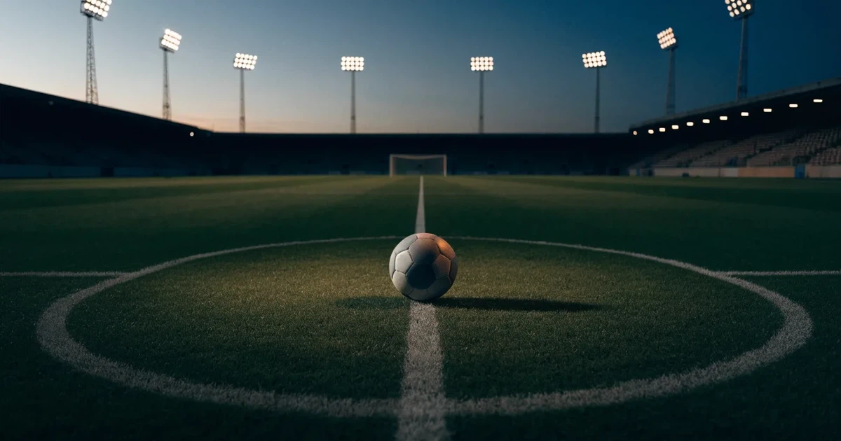 Ein Fußball liegt auf dem Anstoßpunkt eines leeren Rasenplatzes, im Hintergrund eine Tribüne und Flutlichtmasten in der Abenddämmerung.