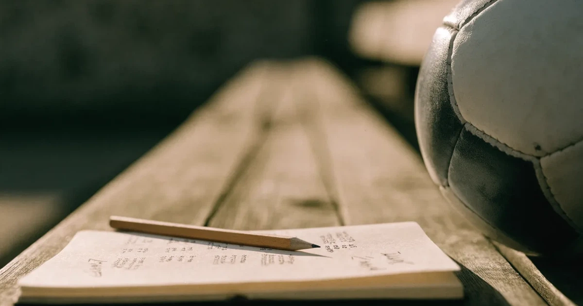 Ein Notizbuch mit handschriftlichen Zahlenkolonnen und ein Bleistift liegen neben einem Fußball auf einer Holzbank.