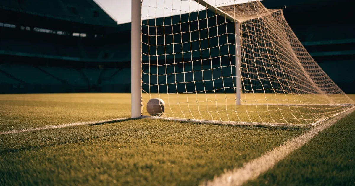 Ein Fußballtor mit Netz im Gegenlicht auf einem leeren Platz am Abend, davor ein Ball auf dem Rasen.