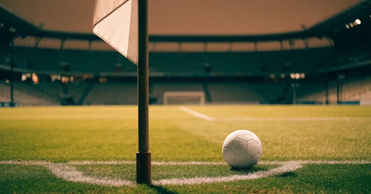 Ein Fußball liegt am Viertelkreis an der Eckfahne eines Rasenplatzes, im Hintergrund leere Tribünen in warmem Abendlicht.