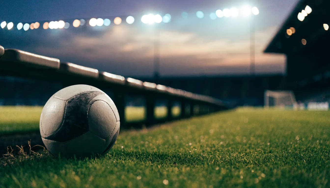 Ein Fußball liegt am Spielfeldrand neben einer leeren Ersatzbank, das Flutlicht über dem Platz leuchtet in der Dämmerung.
