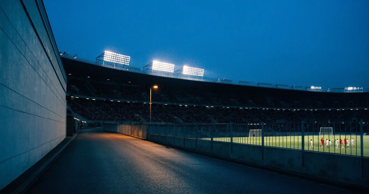 Ein hell erleuchtetes Fußballstadion bei Nacht aus der Ferne gesehen, über dem Dach ein klarer Himmel, davor eine leere Straße.