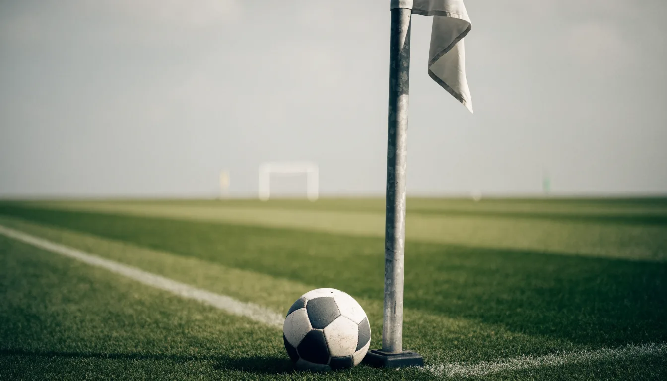 Ein Fußball liegt neben einer Eckfahne auf kurz gemähtem Rasen unter bewölktem Himmel.